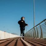 A person wearing a face mask runs alone on an urban bridge pathway under a clear blue sky.