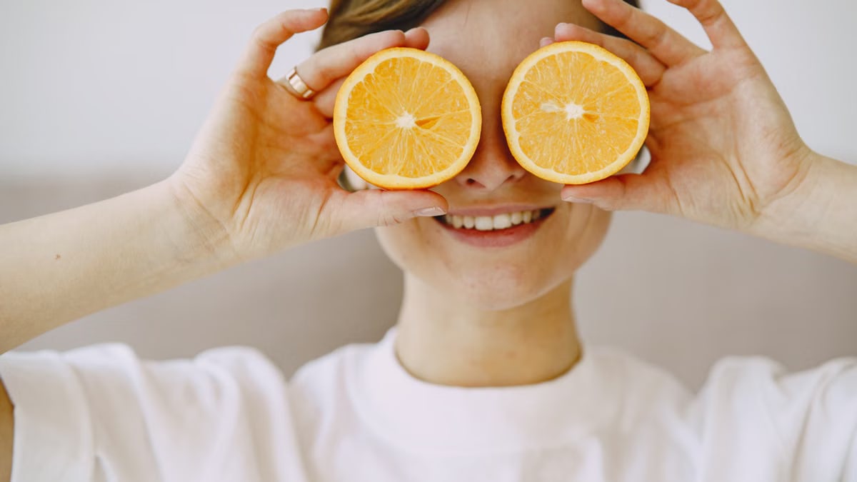 Crop positive female covering eyes with fresh slices of orange smiling and looking forward on blurred background